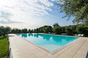 A rectangular swimming pool with a tiled deck, surrounded by greenery and trees.