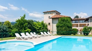 A swimming pool with white lounge chairs, a building with a tiled roof, and greenery in the background. - Apartment in Salò with Pool & Olive Groves (Cunettone)