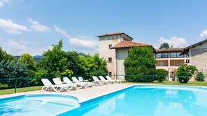A swimming pool with lounge chairs, a building with a tiled roof, and greenery in the background. - Modern Salo Flat with Pool (Cunettone)