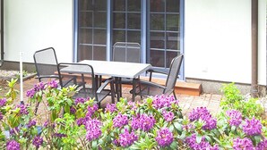 Un bâtiment avec un toit recouvert de tuiles, une fenêtre ornée d’une cloche et un espace extérieur aménagé avec des fleurs violettes.