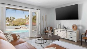 A modern living room with a pool view, a flat-screen TV, and a white cabinet. - Hot Tub, Pool, Firepit, Bball, Foosball, Hammocks (Yucca Valley)