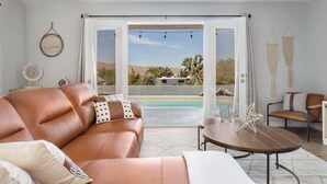 A modern living room with a leather sofa, a round coffee table, and a view of a pool and desert landscape through large glass doors. - Hot Tub, Pool, Firepit, Bball, Foosball, Hammocks (Yucca Valley)