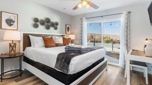A modern bedroom with a large bed, a ceiling fan, and a view of the desert landscape. - Hot Tub, Pool, Firepit, Bball, Foosball, Hammocks (Yucca Valley)