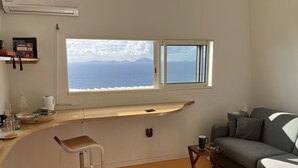 A modern living room with a large window overlooking the sea, a grey sofa, a small table, and a bar stool.