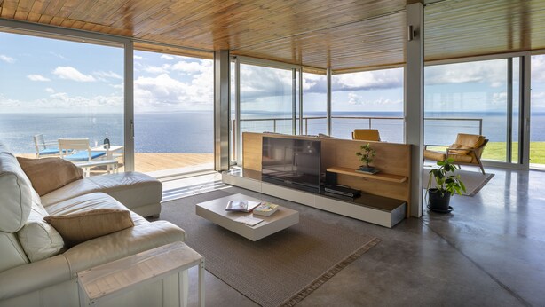 A spacious living room with a large window overlooking the sea, a modern sofa, a coffee table, and a TV stand.