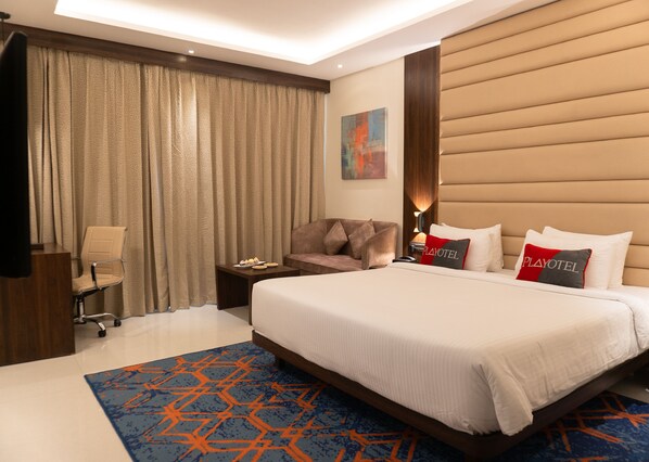 A hotel room with a large bed, a desk, a chair, a nightstand, and a window with curtains.