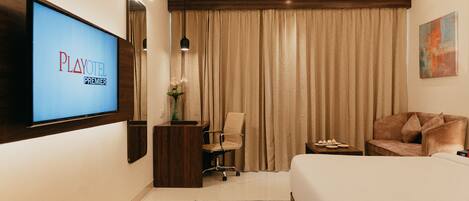 Habitación de hotel con cama, escritorio, silla, televisor mostrando 'PLAYOTEL PREMIER', un florero con flores y un cuadro en la pared.