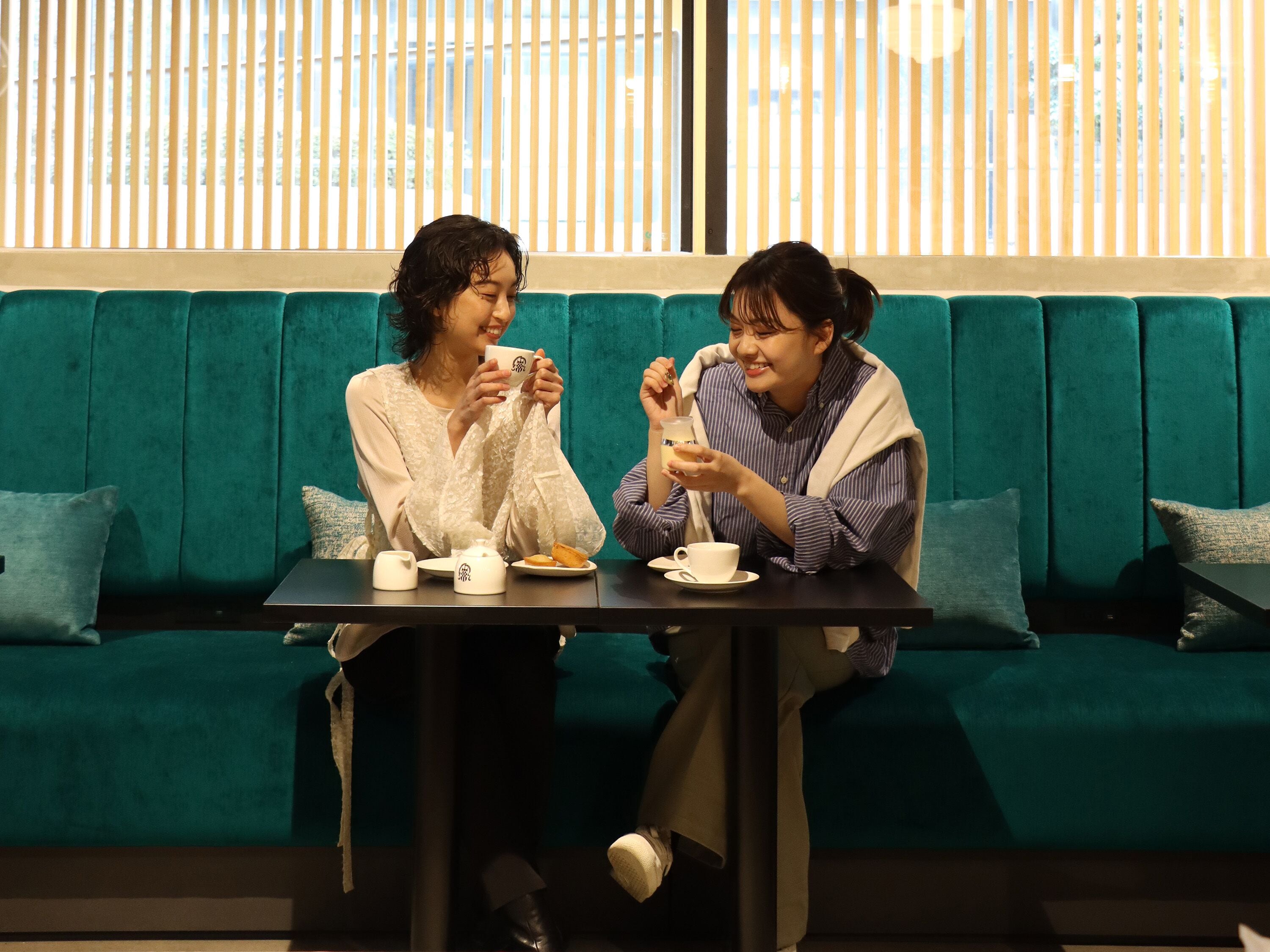Dos personas sentadas en una mesa, con un sofá verde y una ventana con persianas al fondo.