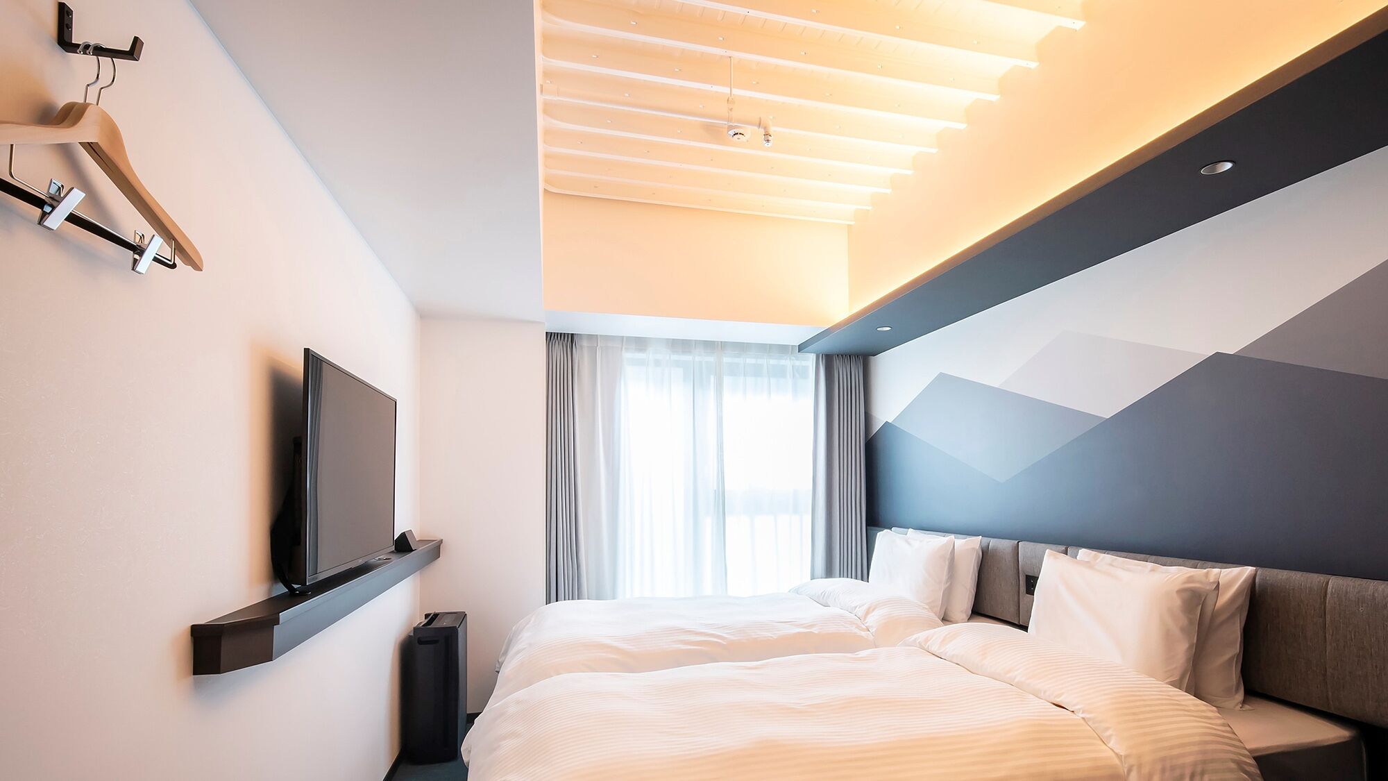 A modern hotel room with a bed, a flat-screen TV mounted on the wall, and a ceiling light fixture.