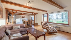 A spacious living room with a wooden ceiling, a large window with a view of trees, a brown leather sofa, a wooden coffee table, and a brown armchair. - Vacation home de Maade for up to 6 people (Enge-Sande)