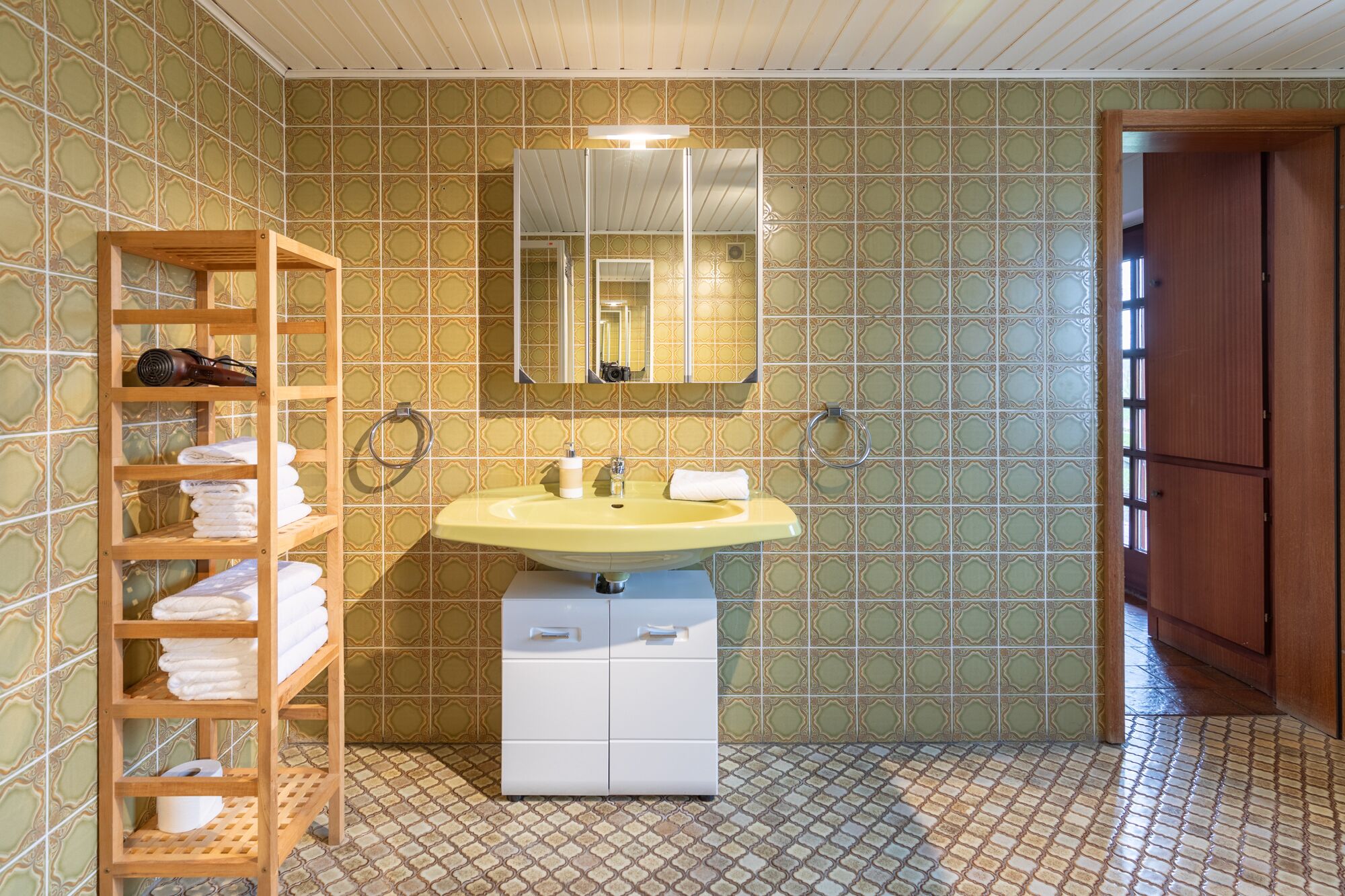 A bathroom with a yellow sink, wooden shelves with towels, a mirror, and patterned tiles.