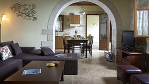 Una sala de estar acogedora con sofá, mesa de centro y comedor con mesa y sillas. Se aprecia un arco de piedra que da a una cocina y un televisor en una esquina.
