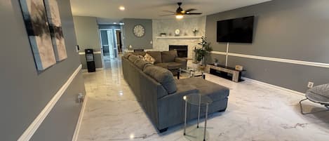 A modern living room with a sectional sofa, a flat-screen TV, a fireplace, and a ceiling fan.