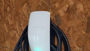 A Tesla charging station with a white charging unit and a blue indicator light, surrounded by coiled black charging cables, set against a wooden background.
