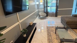 A modern living room with a flat-screen TV mounted on the wall, a black entertainment unit, a glass coffee table, a brown sofa, and a patterned rug. - Beautifully design house for relaxation (Warner Robins)