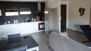 A modern kitchen with white cabinetry, a built-in oven, and a refrigerator. There is a dining table with chairs and a grey sofa in the living area. - Superb villa, 5km from Solenzara. Air-conditioning, swimming pool, fenced grounds. (SOLARO)