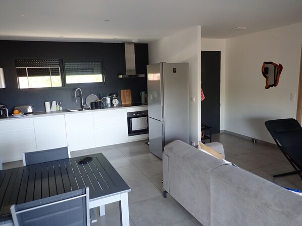 A modern kitchen with white cabinetry, a built-in oven, and a refrigerator. There is a dining table with chairs and a grey sofa in the living area.
