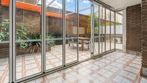 Ein überdachter Patio-Bereich mit Glaswänden, einem gefliesten Boden, einem Holztisch und Stühlen.