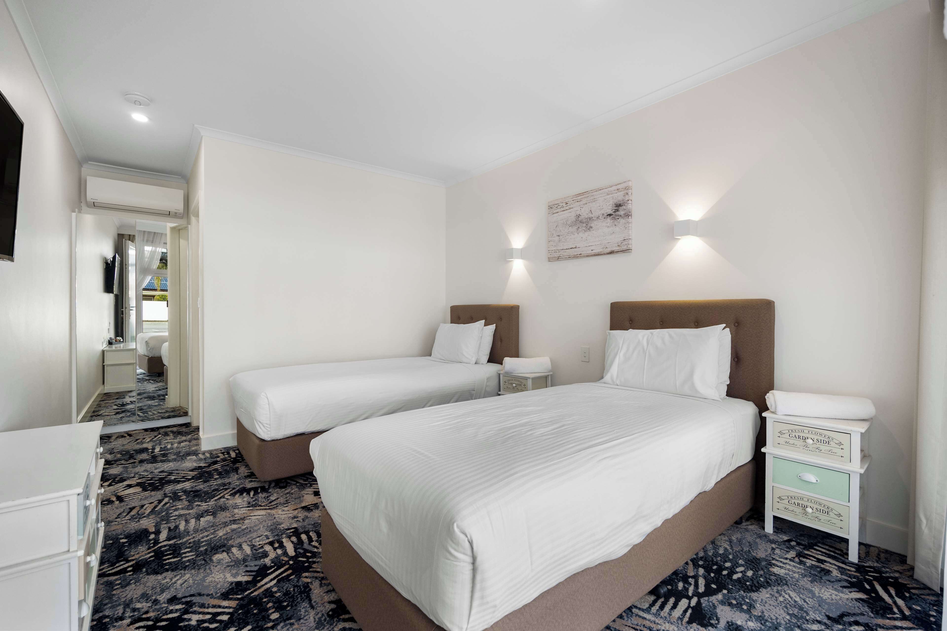 A hotel room with two beds, a dresser, a nightstand with a lamp, and a carpeted floor.