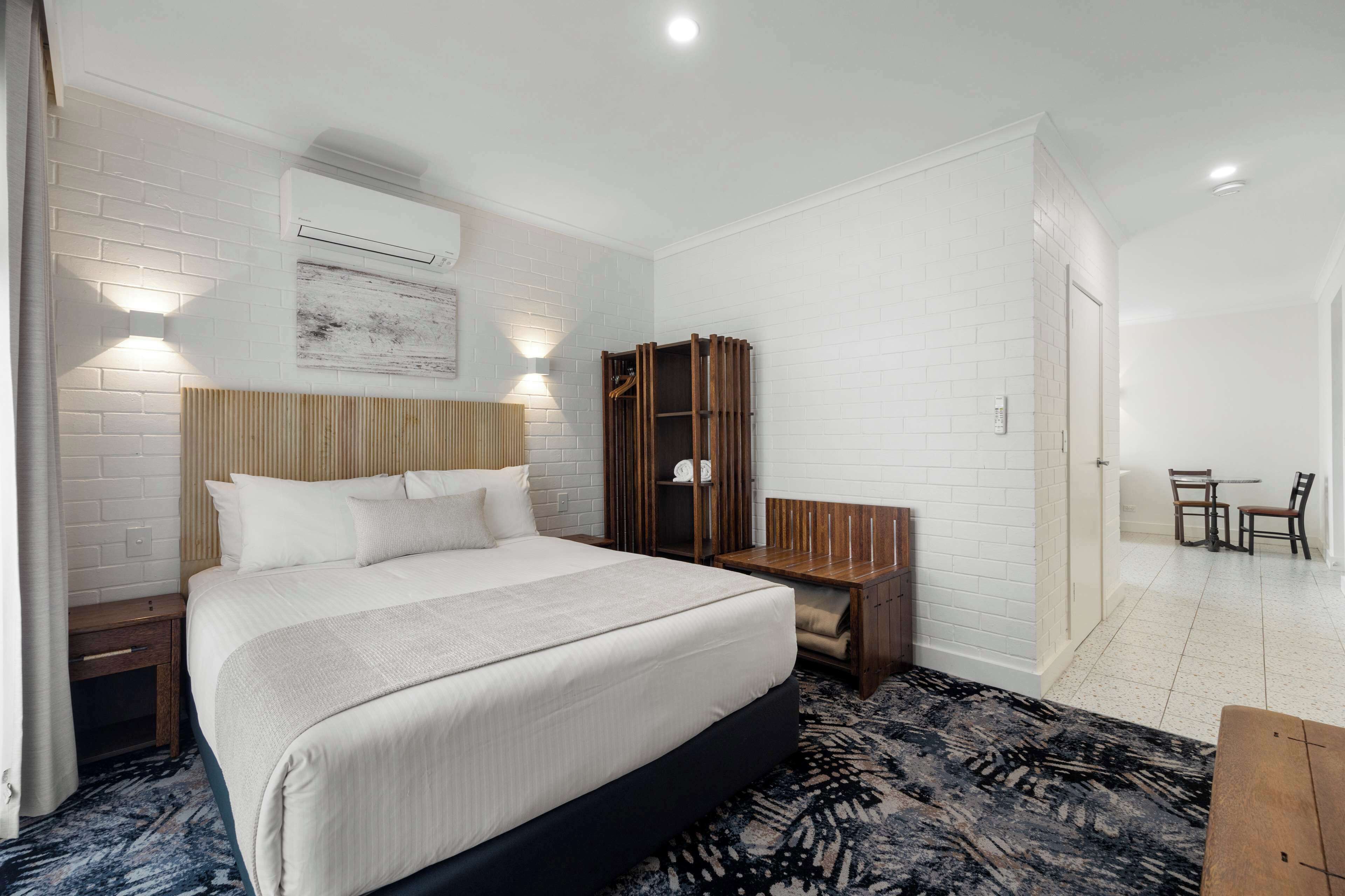Habitación de hotel con una cama grande, una mesita de noche de madera, un armario y una pequeña mesa con sillas.