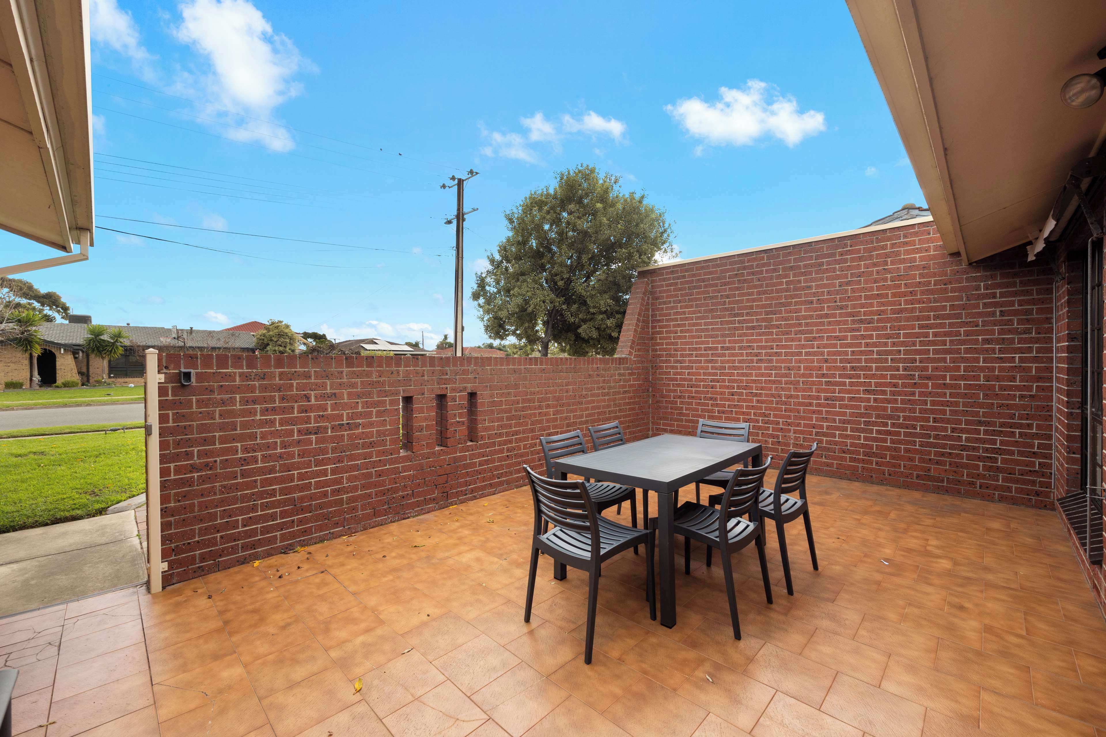 Une terrasse avec un mur en briques, une table avec des chaises et une vue sur un arbre et la rue.