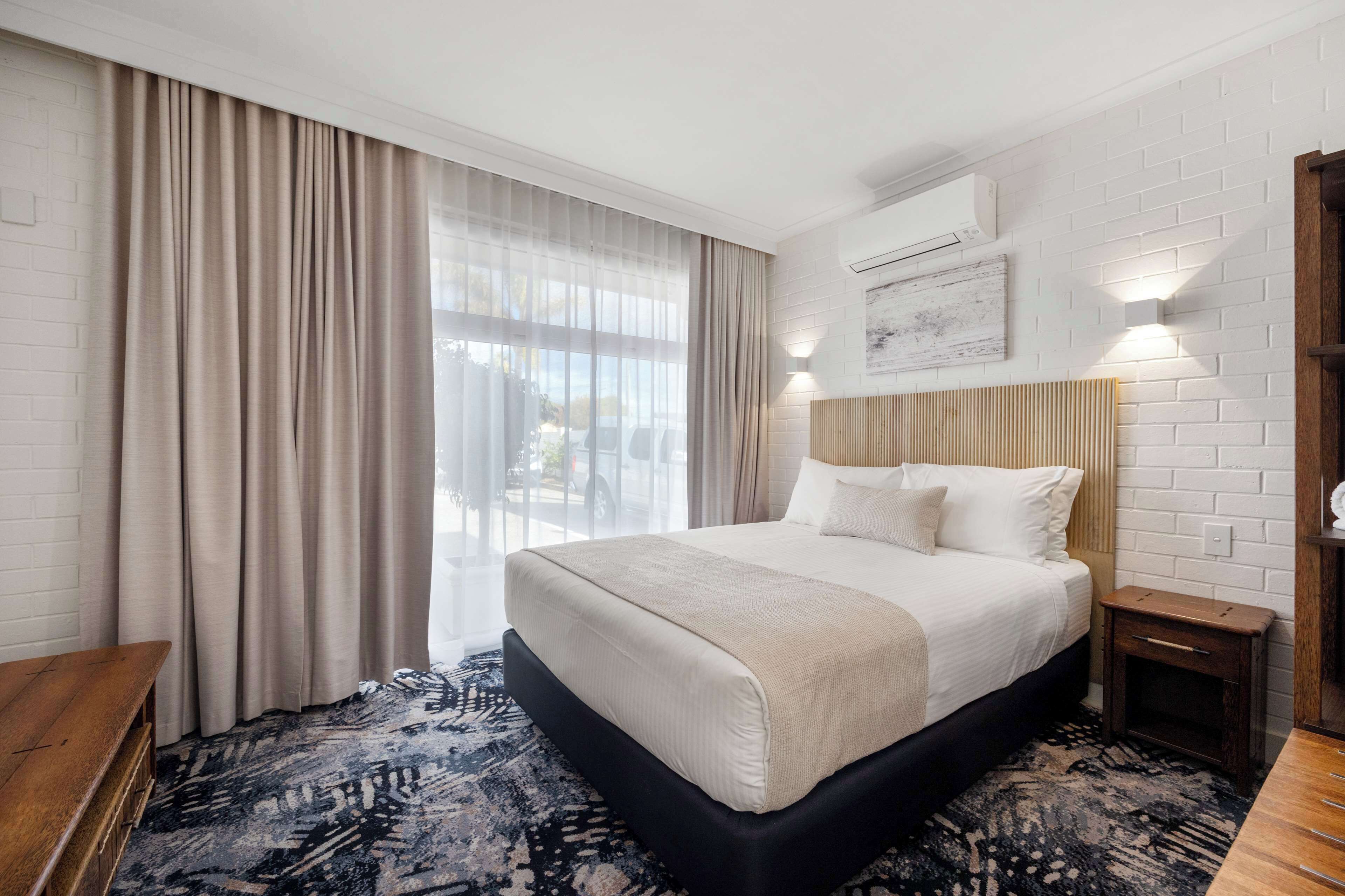 A hotel room with a large bed, a wooden bench, a patterned rug, and a view of the outdoors through sheer curtains.