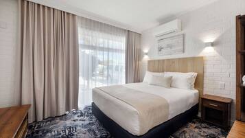 A hotel room with a large bed, a wooden bench, a patterned rug, and a view of the outdoors through sheer curtains.