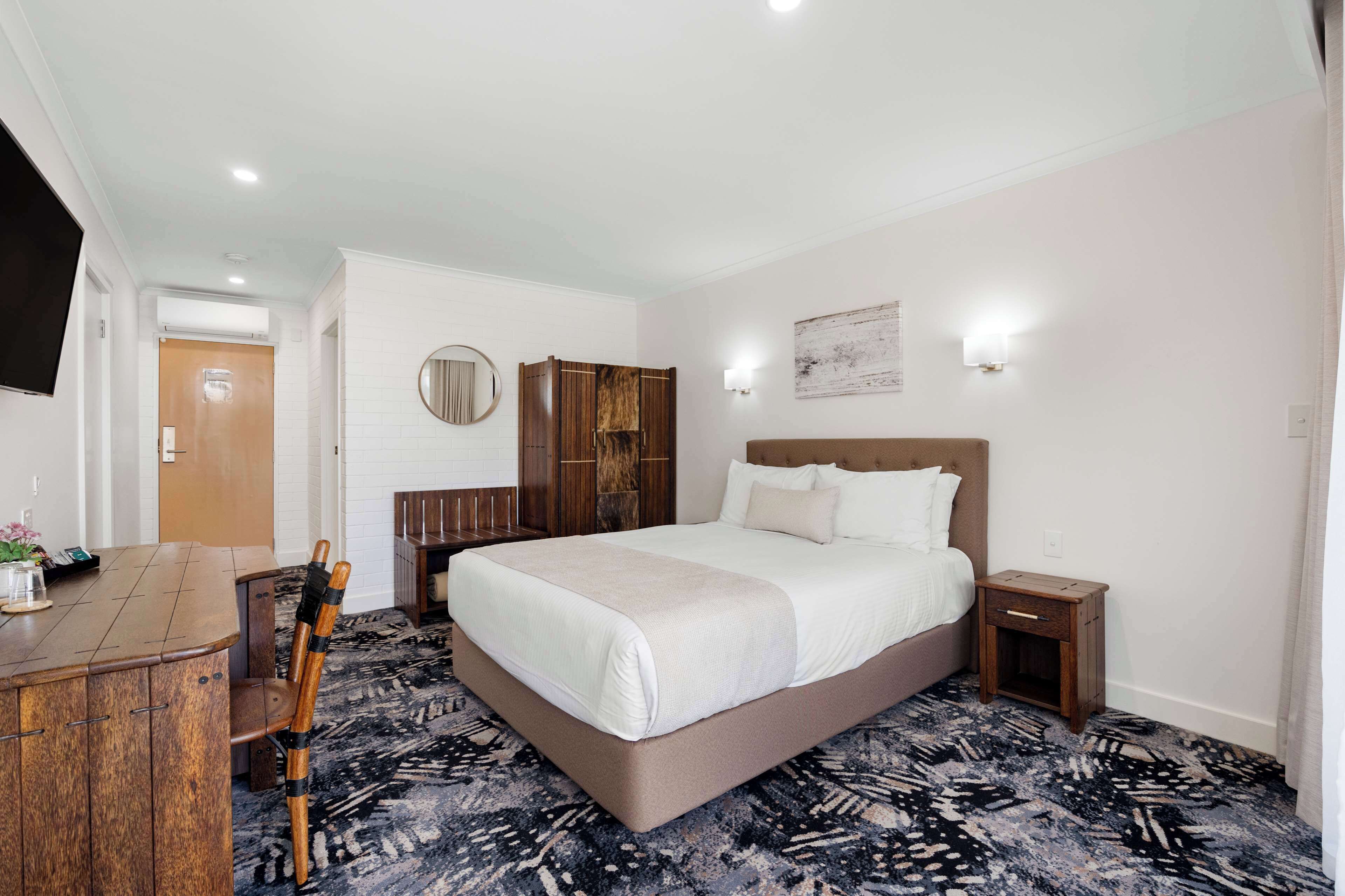 A hotel room with a large bed, a wooden desk, a chair, a wardrobe, and a round mirror on the wall.