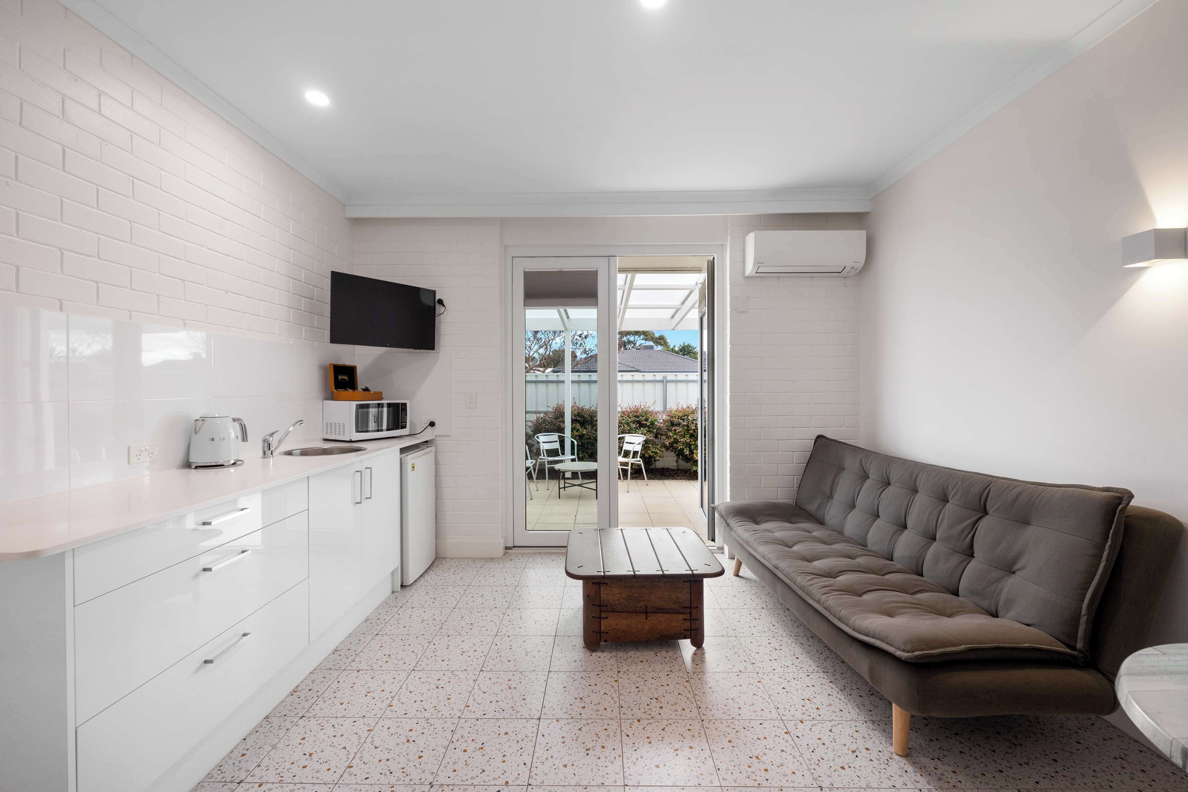 A compact kitchen with white cabinetry, a microwave, and a kettle. There's a wooden table and a grey sofa in the living area, with a glass door leading outside.