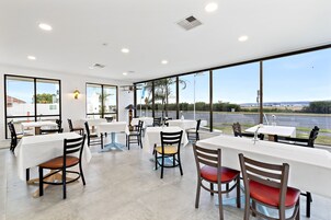 A spacious indoor dining area with large windows offering a view of a road and greenery. Tables are set with white tablecloths and chairs.