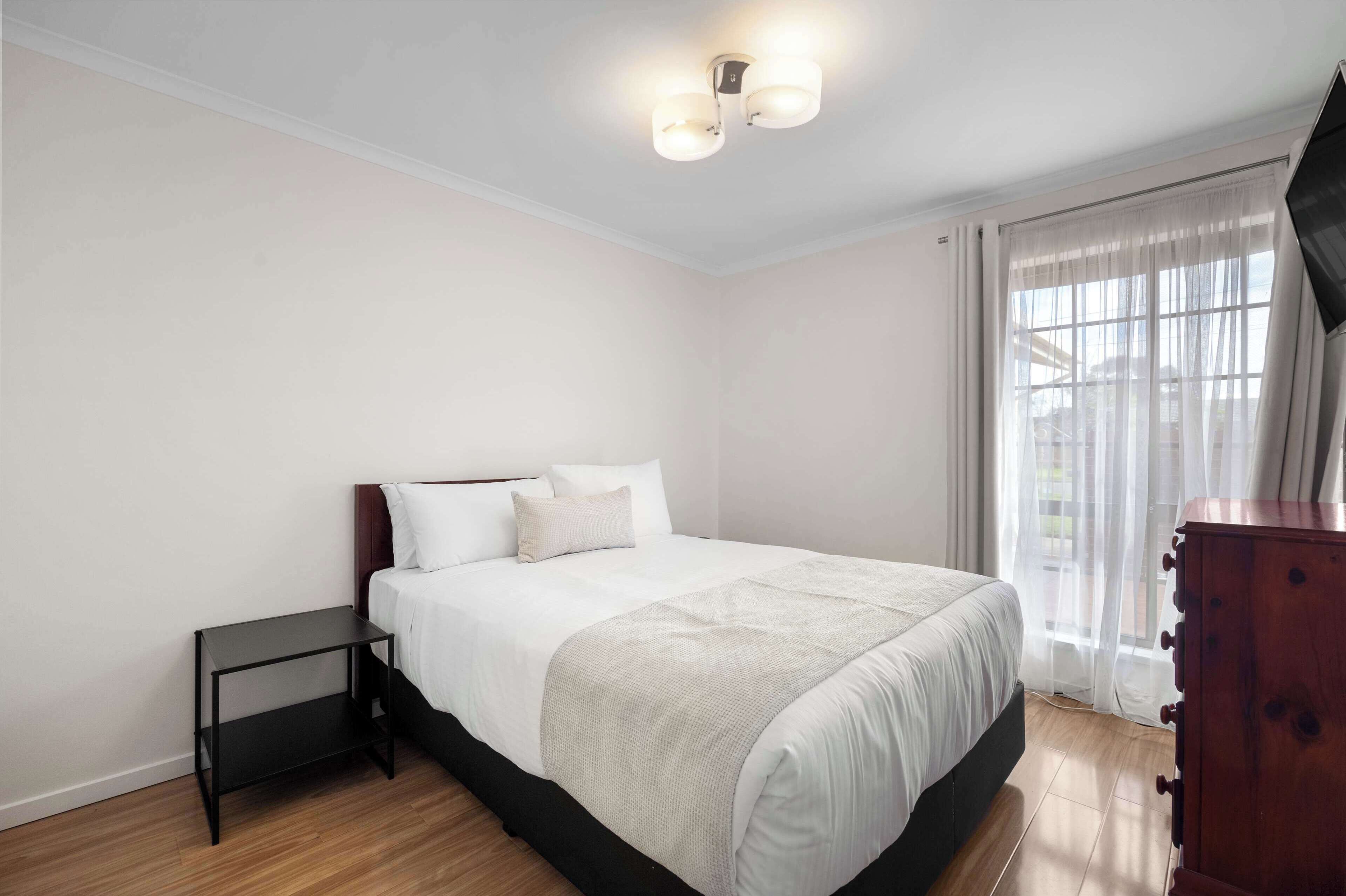 Un dormitorio con cama, mesita de noche, cómoda de madera y ventana con cortinas translúcidas.