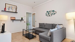 Un salón con un sofá gris, una mesa de centro de cristal, una mesita auxiliar blanca, un televisor sobre un mueble, un cuadro en una estantería y un adorno en la pared.