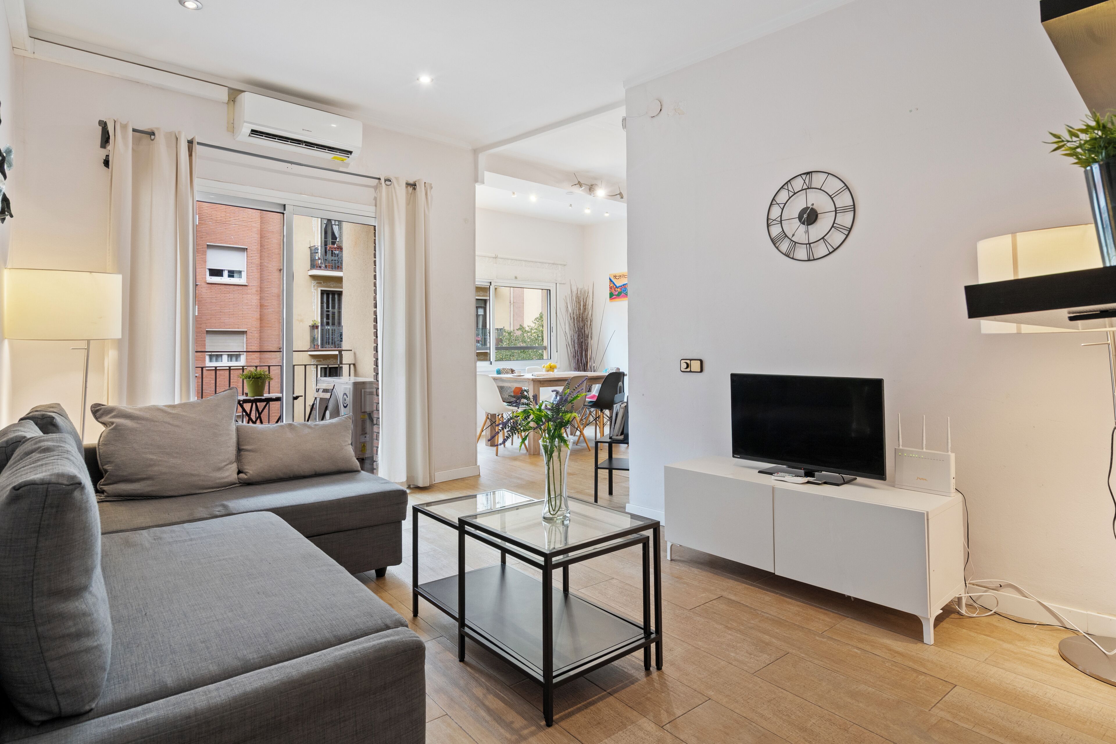 A modern living room with a sofa, a glass coffee table, a TV on a white cabinet, and a clock on the wall.