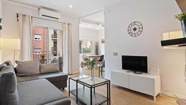 Una sala de estar moderna con un sofá, una mesa de centro de vidrio, un televisor sobre un mueble blanco y un reloj en la pared.