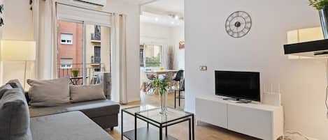 A modern living room with a sofa, a glass coffee table, a TV on a white cabinet, and a clock on the wall.
