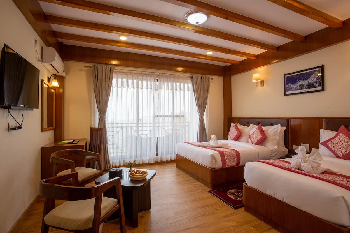 A hotel room with wooden beams, two beds with red and white bedding, a desk with a chair, a TV, and a window with sheer curtains.