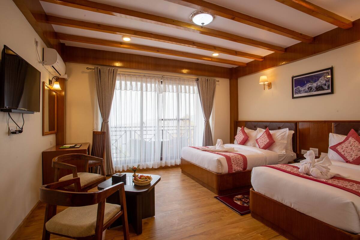 A hotel room with wooden beams, a television, two beds with red pillows, a desk with a chair, and a window with curtains.