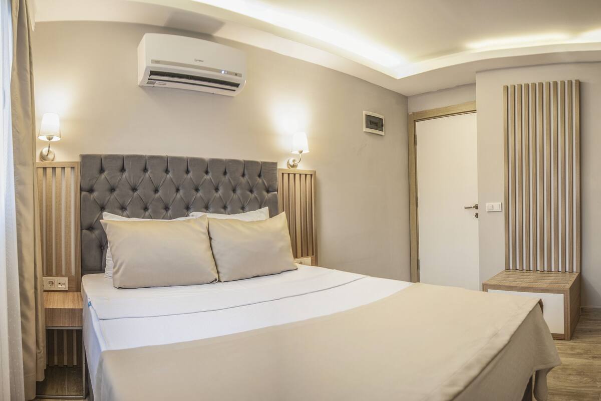 A hotel room with a bed, bedside lamps, an air conditioner, a television, and a wooden desk.