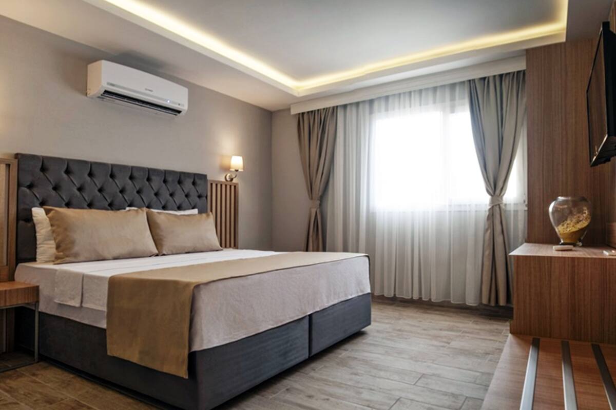 A modern hotel room with a large bed, a flat-screen TV, an air conditioning unit, and a wooden nightstand.