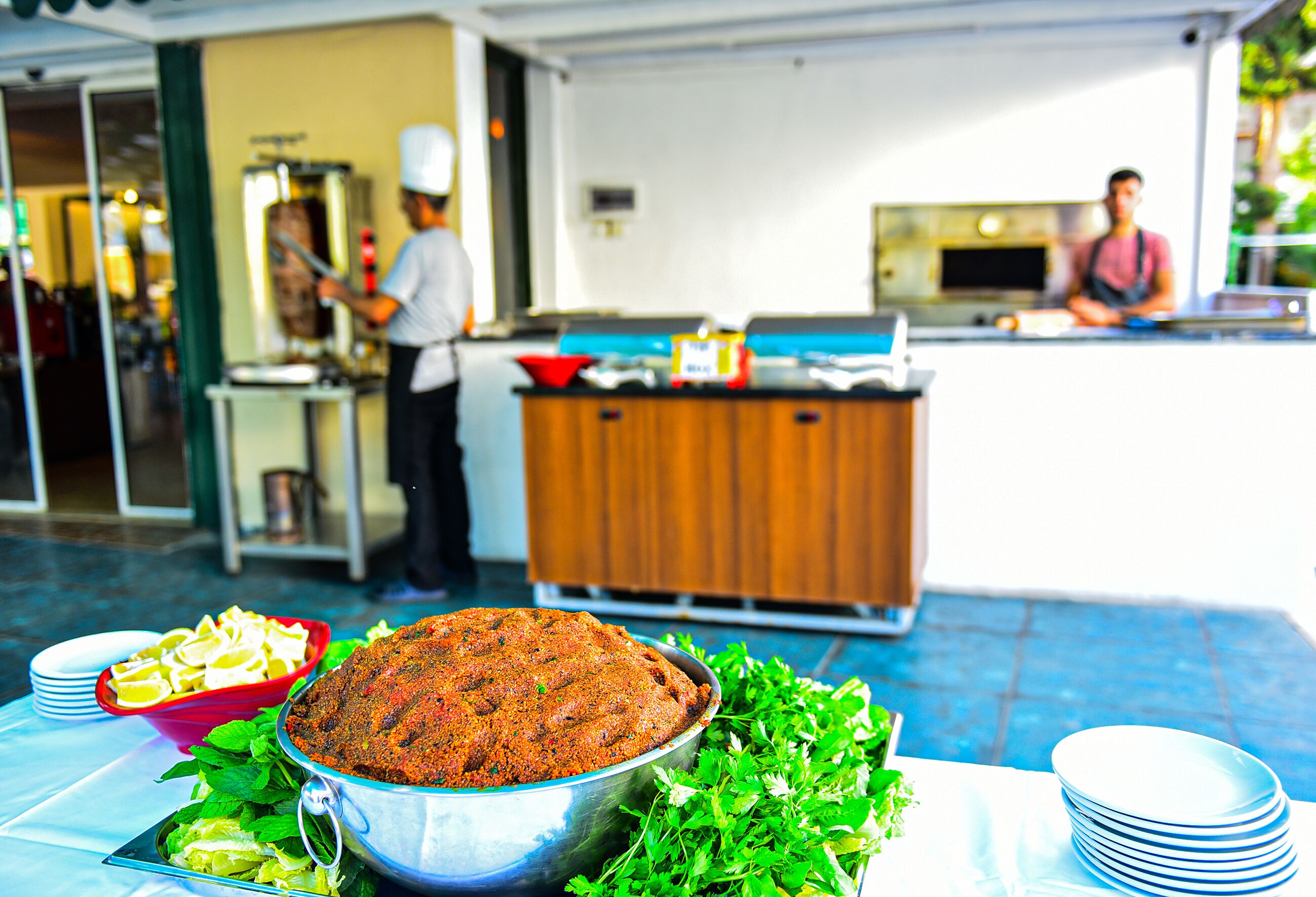 A buffet table with a large metal bowl of food, a red bowl of salad, and plates. In the background, there are two people working behind a counter.