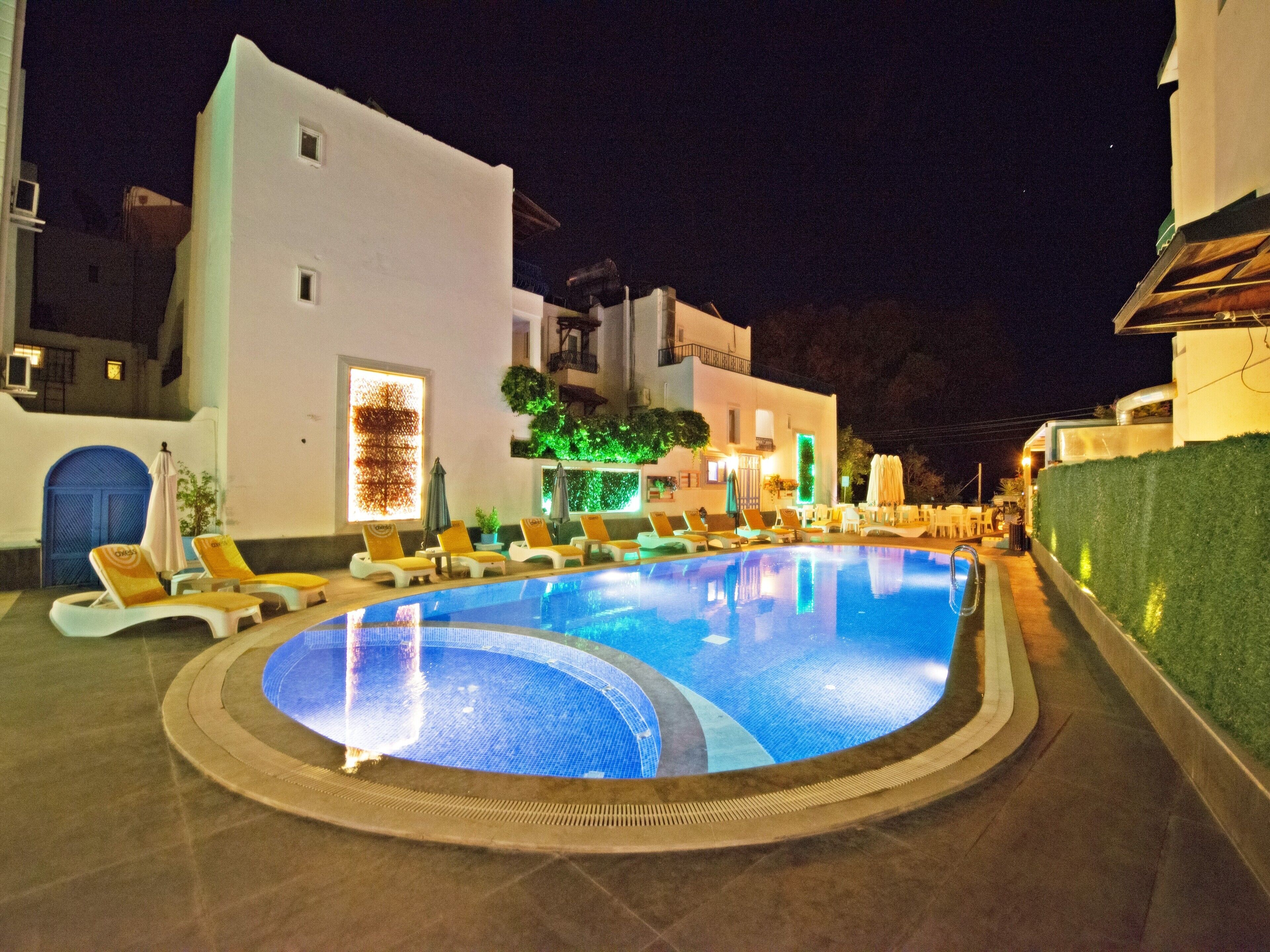 Ein Schwimmbeckenbereich bei Nacht mit Liegestühlen und einem Gebäude mit heller Fassade.