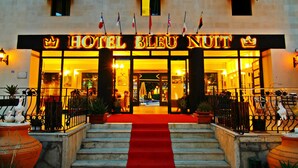 De ingang van Hotel Bleu Nuit, met een rode loper die naar de deuren leidt, vlaggen en een stenen gevel.