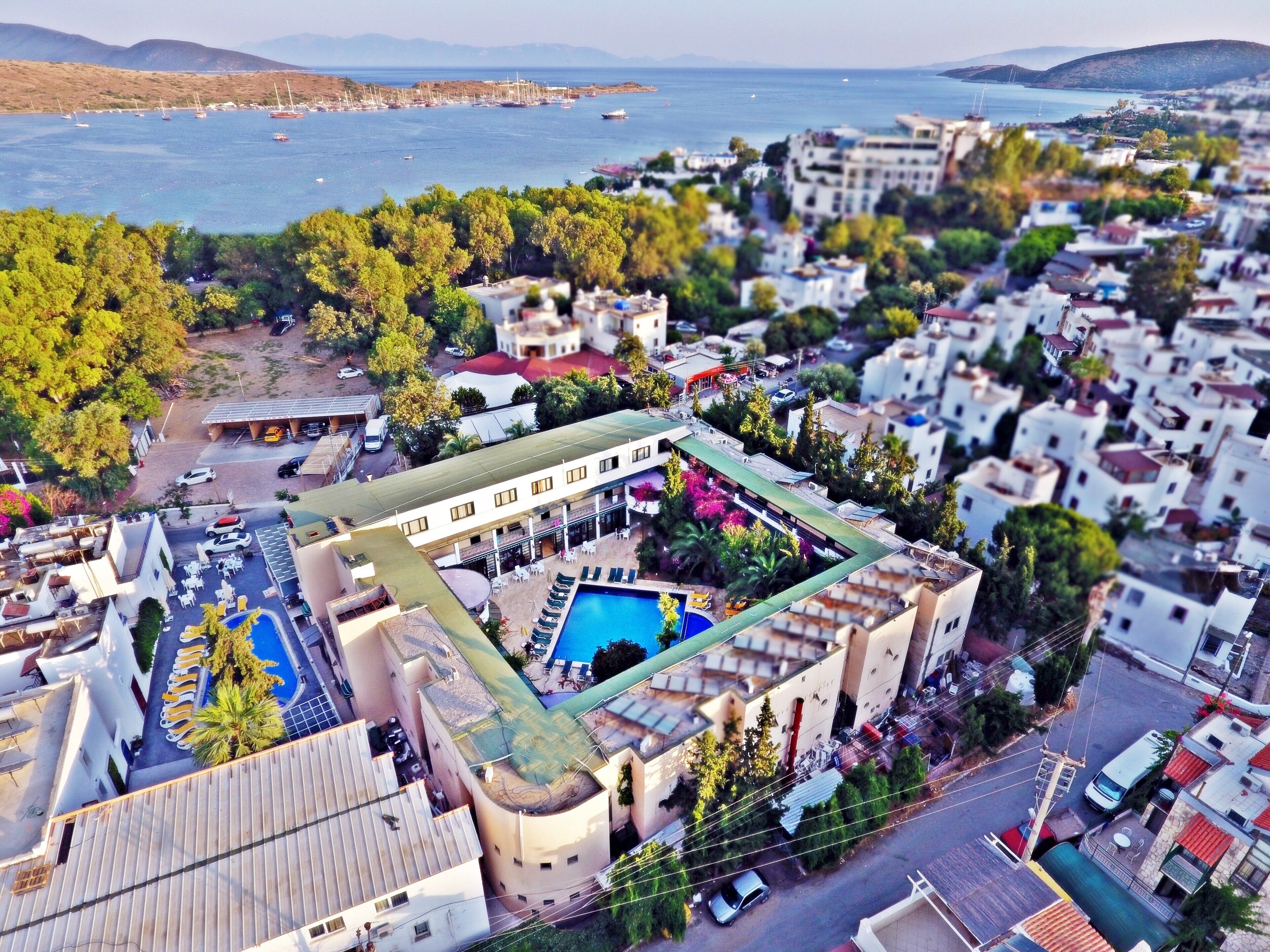 An aerial view of a coastal town with a prominent hotel featuring a rooftop pool, surrounded by lush greenery and a marina with boats.