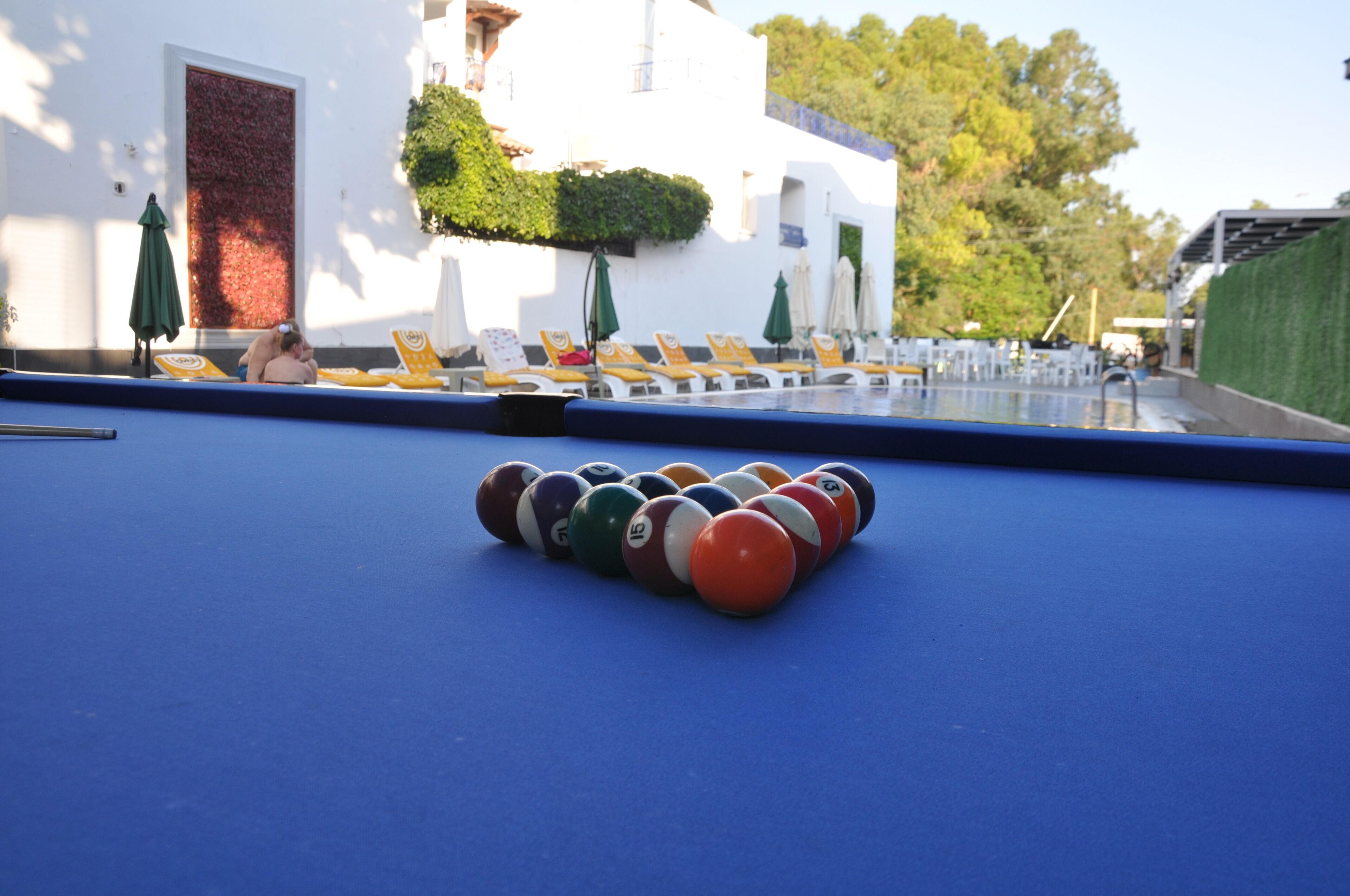 A pool table with numbered balls in front of a building with outdoor seating and umbrellas.