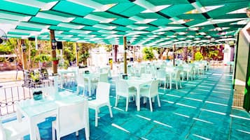 An outdoor dining area with white tables and chairs, covered by a green and white patterned canopy.
