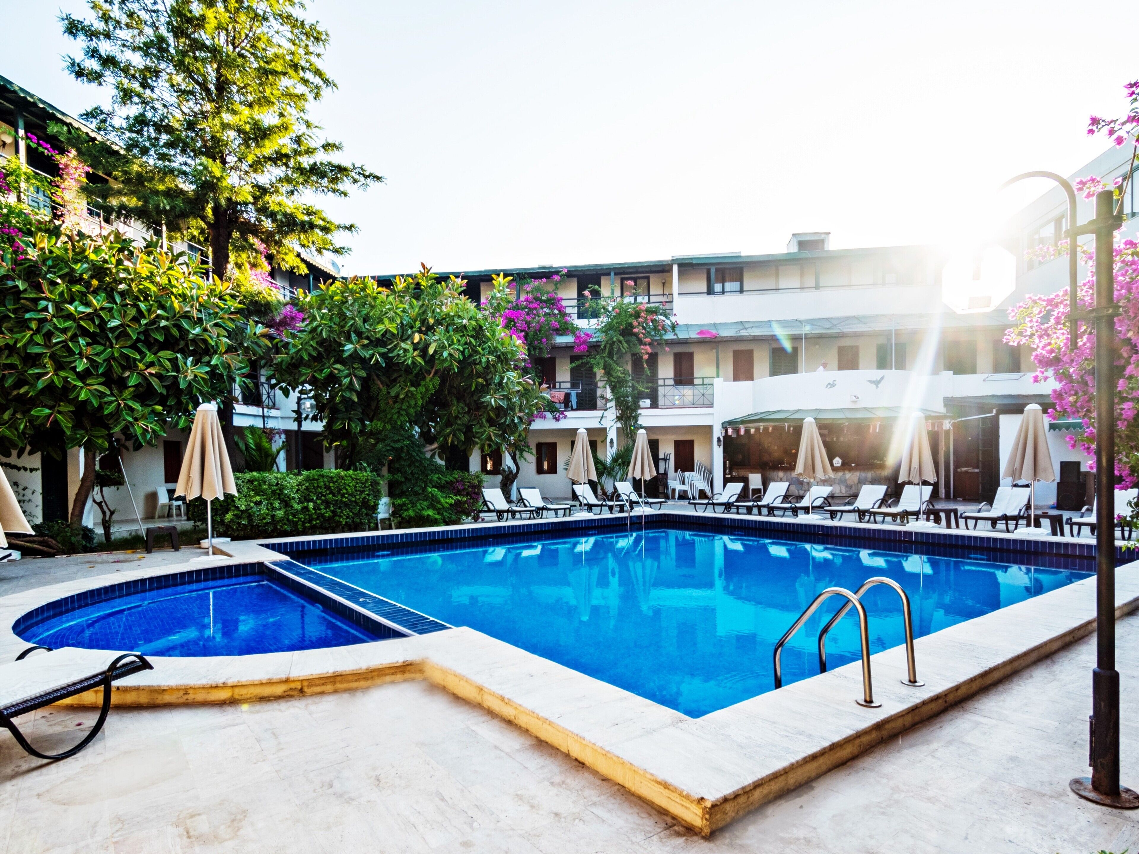 Une piscine entourée de verdure, avec des chaises longues et des parasols, et un immeuble à plusieurs étages en arrière-plan.