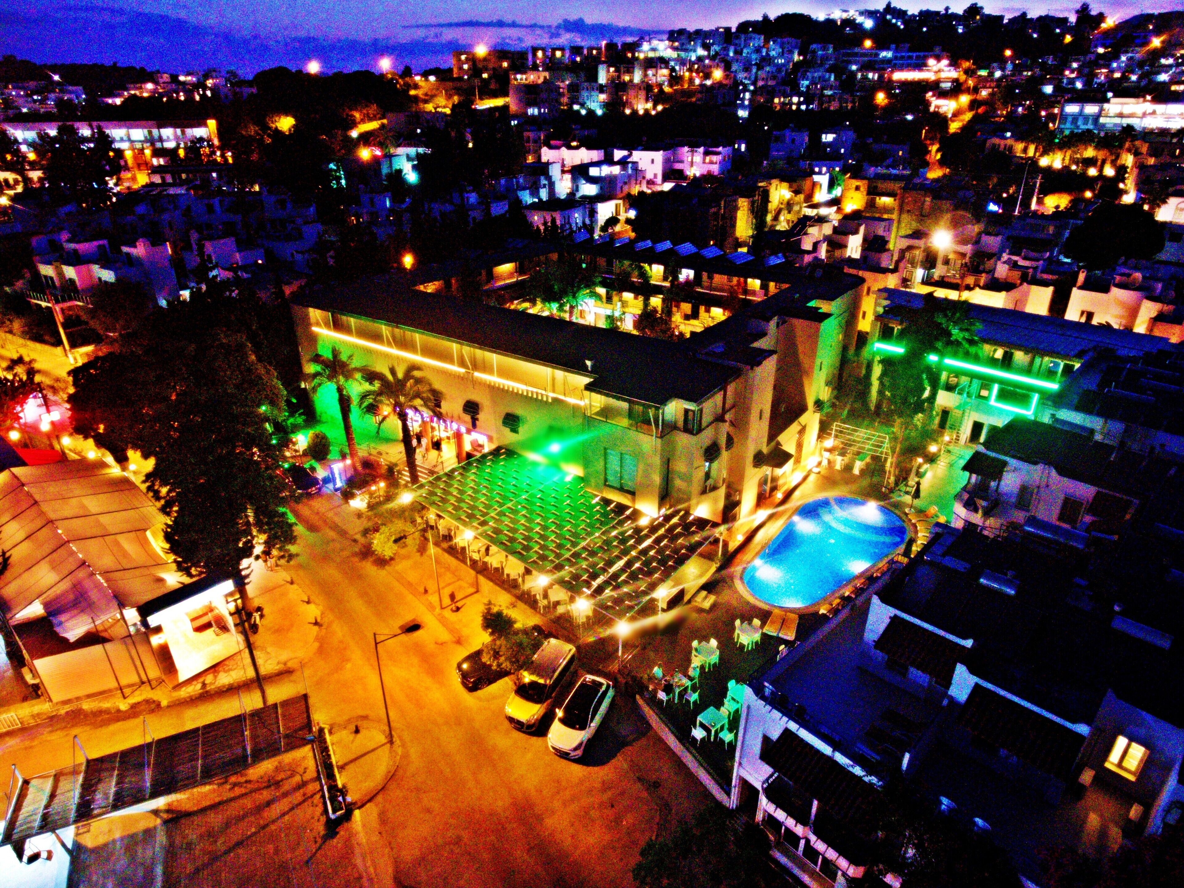 An aerial view of a hotel with a swimming pool, outdoor seating area, and a well-lit facade.