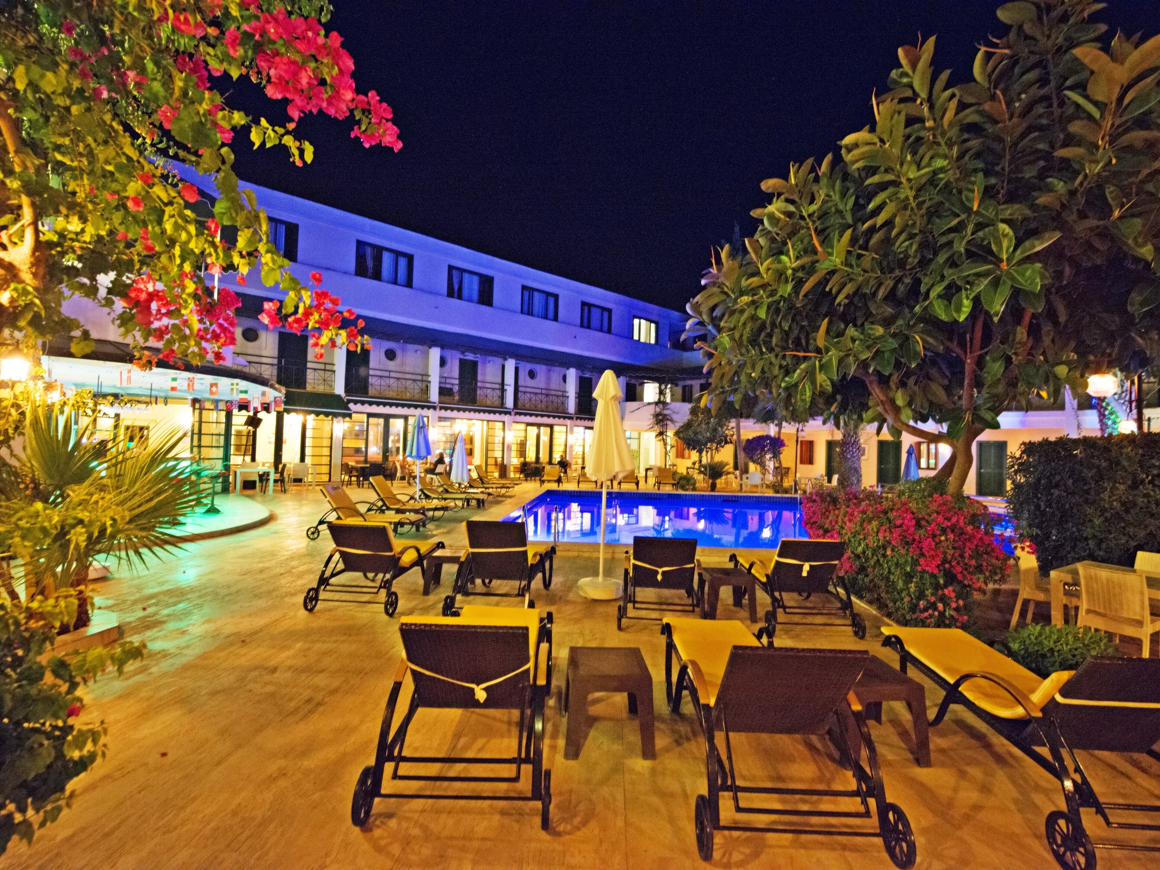 A poolside area with lounge chairs and a swimming pool, surrounded by greenery and a building with multiple windows.