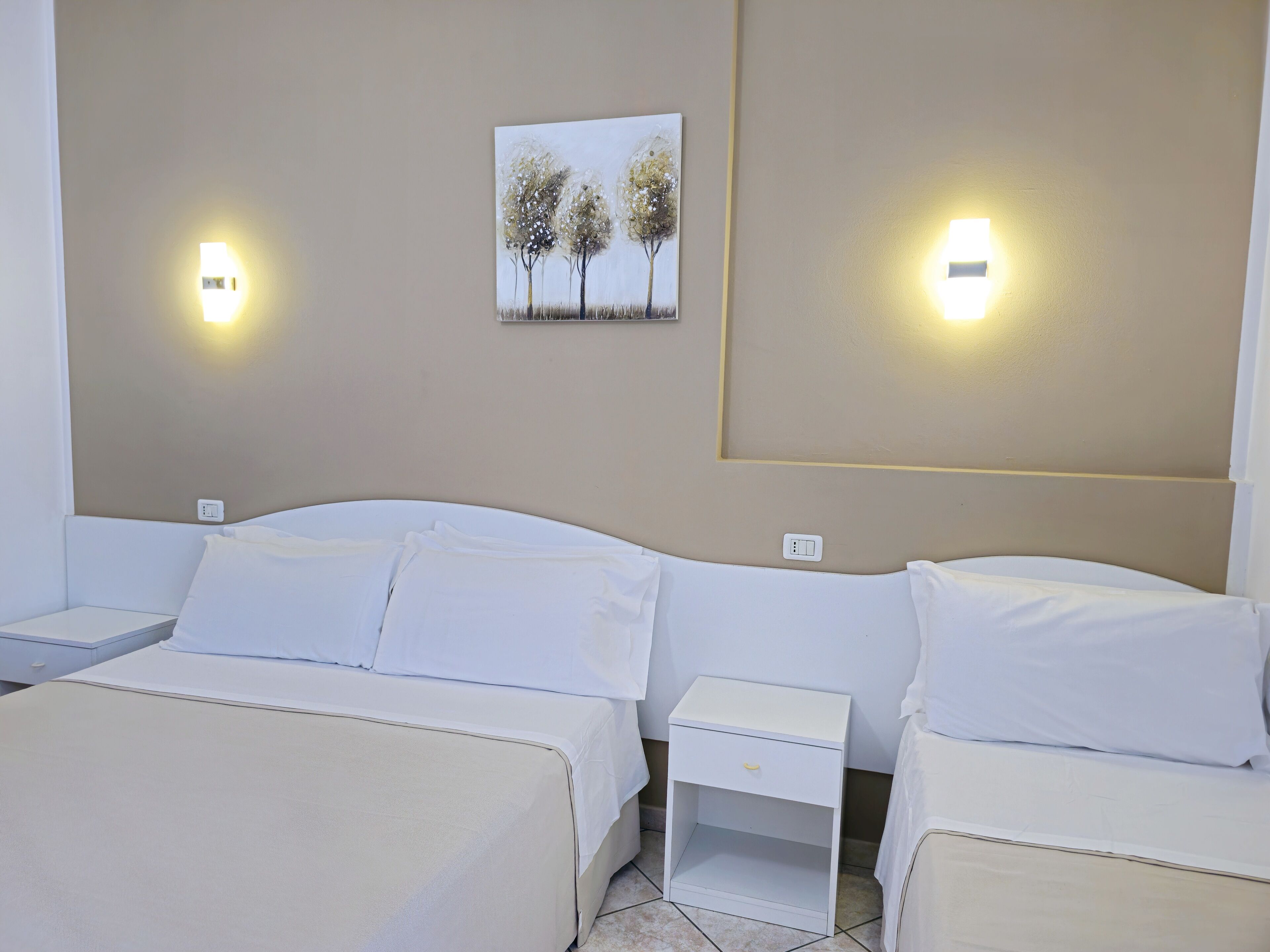 A hotel room with two beds, white bedding, a nightstand with a drawer, and a wall-mounted painting of trees.
