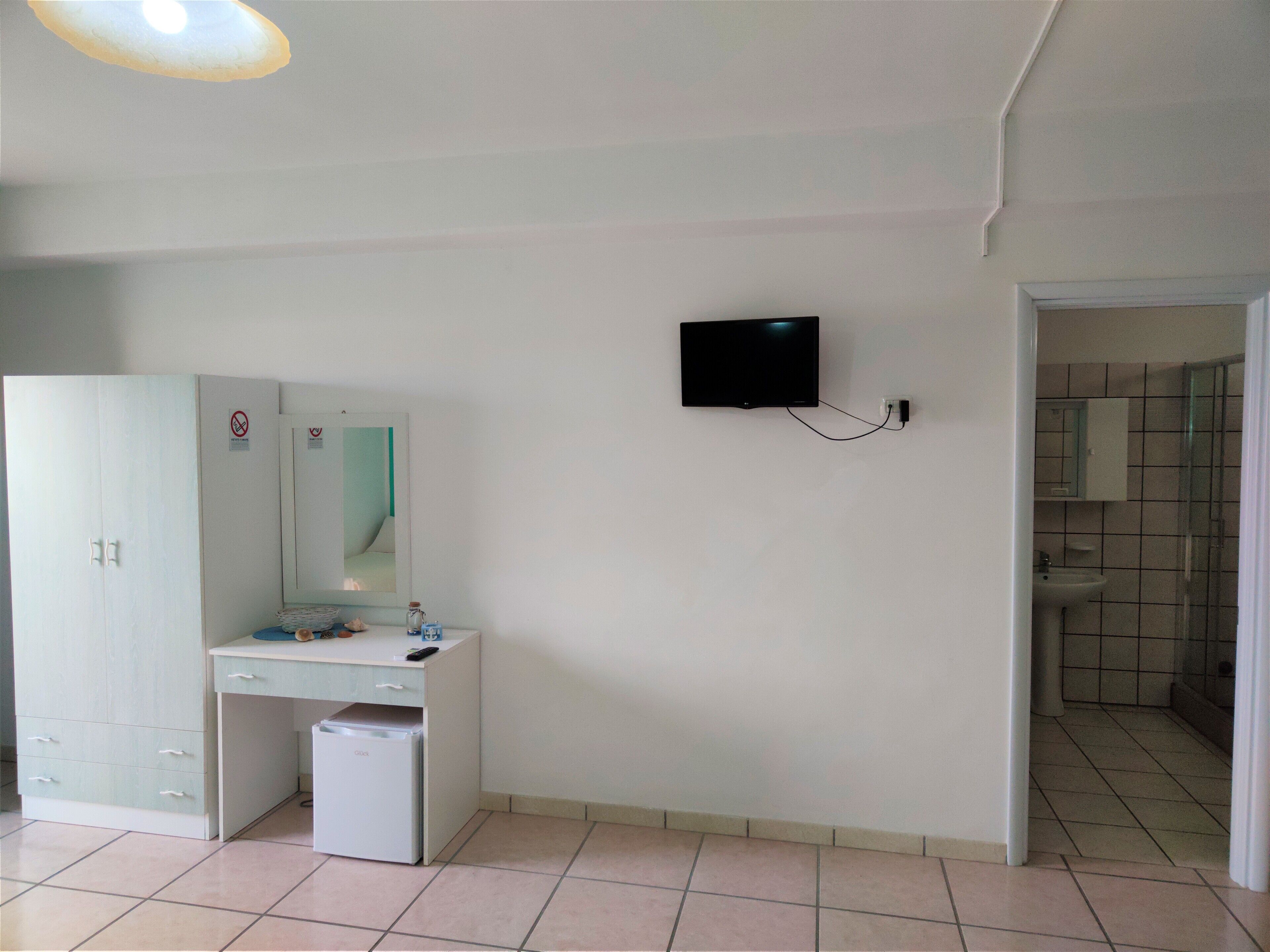 A room with a white wardrobe, a desk with a mirror, a small fridge, and a TV mounted on the wall.